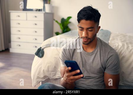 Giovane uomo biraciale in abbigliamento casual utilizzando il telefono cellulare in camera da letto a casa Foto Stock