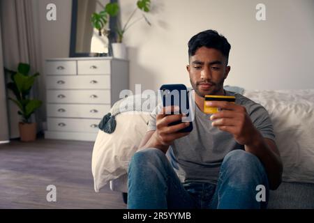 Giovane uomo biraciale in abbigliamento casual utilizzando il telefono cellulare per le banche in camera da letto a casa Foto Stock