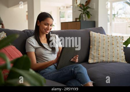 Felice giovane donna biraciale in abbigliamento casual utilizzando il tablet digitale sul divano in soggiorno Foto Stock