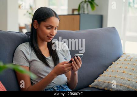 Contented giovane donna biraciale in abbigliamento casual utilizzando il telefono cellulare sul divano in soggiorno Foto Stock