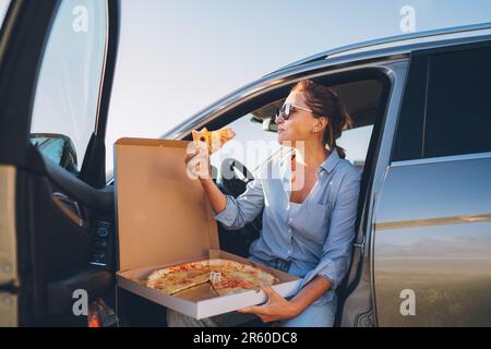 Donna anziana che mangia pizza italiana appena cotta seduto sul sedile dell'autista durante la pausa pranzo e godendosi la luce del tramonto. Auto in viaggio, fast food ea Foto Stock