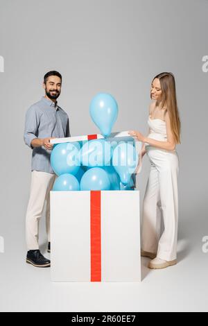 Allegro ed elegante donna incinta apertura grande scatola regalo con  palloncini blu vicino al marito durante la celebrazione e il sesso rivelare  la festa a sorpresa sul grigio b Foto stock 