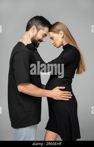 Vista laterale di donna elegante e incinta dai capelli leali in vestito nero che abbraccia marito sorridente e bearded mentre si è in piedi con gli occhi chiusi isolato su gre Foto Stock