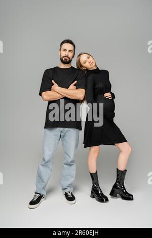 Intera lunghezza di donna in gravidanza elegante in abito nero che tocca la pancia e guardare la macchina fotografica mentre si trova vicino al marito con le braccia incrociate sul dorso grigio Foto Stock