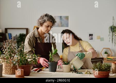 Ragazza con sindrome di Down innaffiare le piante mentre le piantano insieme con fiorista in camera Foto Stock