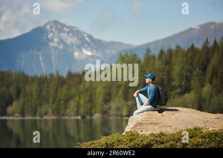 Uomo con zaino seduto sulla roccia e con vista panoramica sul lago di montagna. Giovane viaggiatore da solo nelle Alpi. Foto Stock