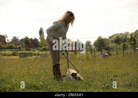 Una donna bionda in piedi in un campo di cavalli raccolta poo Foto Stock