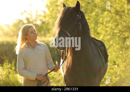 Una donna dai capelli biondi in piedi con un cavallo nero in una serata di sole Foto Stock