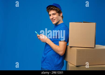 Giovane corriere in uniforme blu sorridente guardare il suo smartphone mentre si appoggia su pacchi scatola di cartone Foto Stock