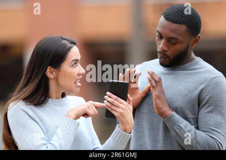 Una donna arrabbiata che scola un uomo scioccato che mostra il contenuto del telefono cellulare per strada Foto Stock