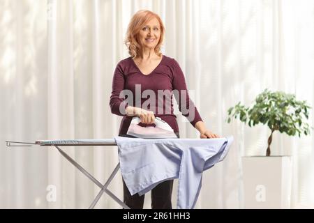 Donna matura che stira una camicia e sorride a casa Foto Stock