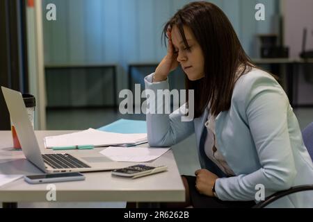 Una giovane donna lavora in un ambiente professionale, seduto alla sua scrivania con il computer portatile, la documentazione e altri materiali davanti a lei Foto Stock