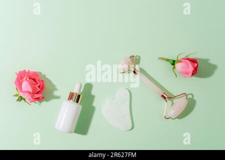 Rullo facciale in cristallo di quarzo rosa e raschietto gua sha, siero facciale o olio cosmetico organico su sfondo verde. Kit massaggio viso per lifting therapy. Foto Stock