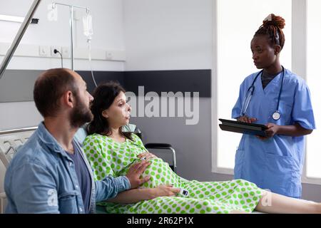 Assistente afroamericano che discute il processo di parto con la donna incinta, preparante la madre per l'ambulatorio nel reparto dell'ospedale. Paziente con gravidanza sdraiato a letto in attesa di medico ginecologo Foto Stock