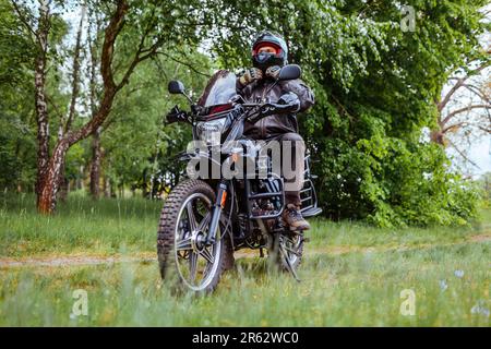 Il biker svezza il casco dopo aver cavalcato il motobike nella foresta estiva. Uomo che indossa abiti in pelle all'aperto. Conducente seduto durante il trasporto. Viaggio estivo Foto Stock