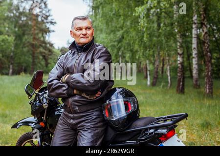 Biker anziano appoggiato su motobike nella foresta estiva. Uomo in pelle vestiti in piedi con le braccia incrociate godendo la natura. dopo il giro Foto Stock
