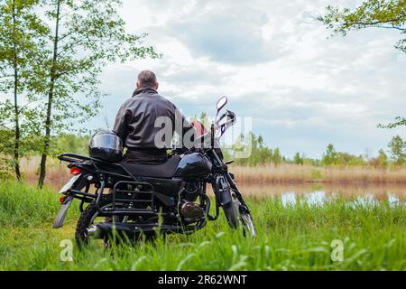 Vista posteriore di un motociclista anziano seduto su una moto all'aperto. Uomo ammirando il paesaggio del fiume rilassante. Il pilota che viaggia godendo la natura Foto Stock