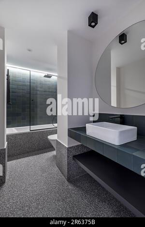 Bagno ristrutturato di recente con pavimenti in terrazzo, piano di appoggio piastrellato, specchio circolare, lavabo in porcellana bianca e vassoio doccia Foto Stock