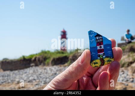 06 giugno 2023, Schleswig-Holstein, Falshöft: Una donna tiene una pietra dipinta trovata sulla spiaggia in aria. Sulla pietra si può vedere il faro di Falshöft, che si erge nell'originale su un tratto di spiaggia nell'omonimo villaggio. Falshöft è l'ingresso al Geltinger Birk. La riserva naturale 'Geltinger Birk' si trova all'uscita del fiordo di Flensburg. Con una superficie totale di 773 ha, è la più grande riserva naturale del distretto Schleswig-Flensburg. Foto: Frank Hammerschmidt/dpa Foto Stock