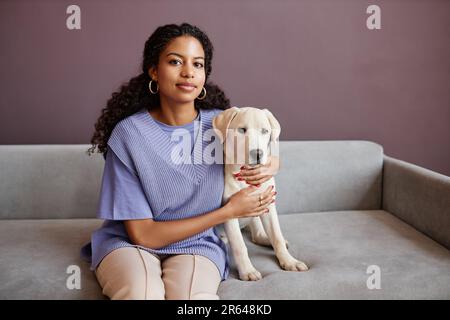 Ritratto minimo di giovane donna nera con cane seduto sul divano e guardando la macchina fotografica in toni viola, spazio copia Foto Stock
