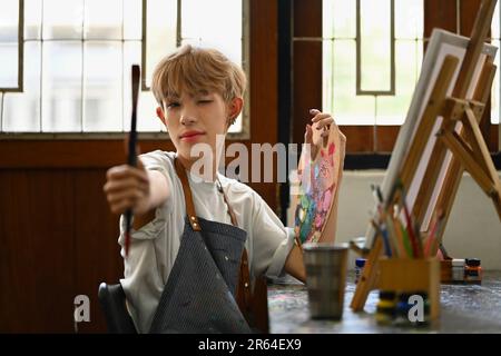 Grazioso uomo gay adolescente che tiene pennelli e tavolozza seduti in studio d'arte. Educazione, hobby, concetto di arte Foto Stock