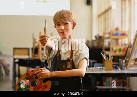 Grazioso uomo gay adolescente che tiene pennelli e tavolozza seduti in studio d'arte. Educazione, hobby, concetto di arte Foto Stock