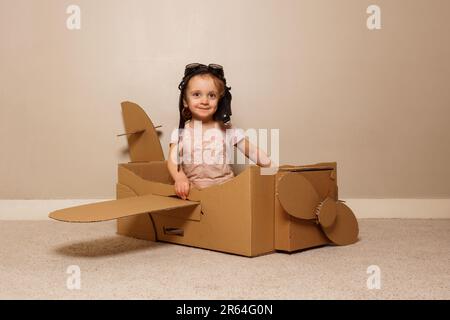La ragazza si siede in un piano di cartone, indossa un cappello, gli occhiali fingono di volare Foto Stock
