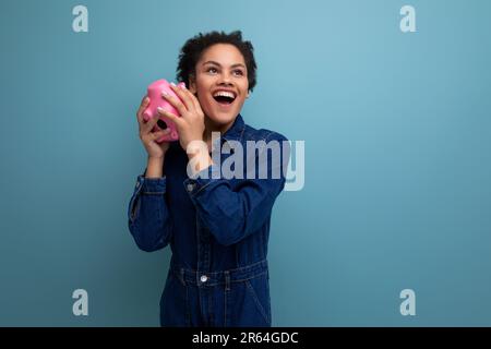 giovane e snella donna brunetta ispanica con capelli ricci morbidi in un tuta di denim blu pensa di spendere i risparmi dalla banca del piggy Foto Stock