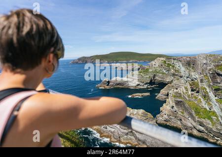 Blonde Woman che guarda alla maestà Cliffside dell'Irlanda, ipnotizzata da Azure Skies Foto Stock