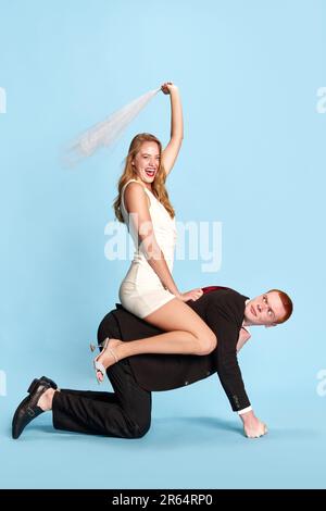 Sposarsi. Giovane donna felice ed eccitata seduta sulla schiena dell'uomo contro lo sfondo blu dello studio. Moglie felice Foto Stock