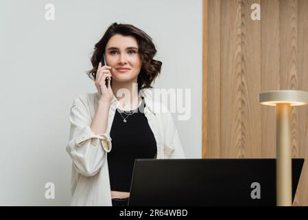 una giovane donna piaciuta in abiti eleganti e informali, con capelli brunette ondulati e tatuaggi che parlano al telefono mentre lavoravano come receptionist alla reception nea Foto Stock