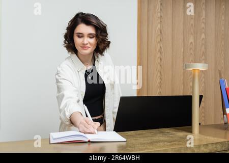 concierge cordiale in abiti eleganti e informali, con scrittura dei capelli in brunette ondulata nel notebook vicino al monitor del computer e alla lampada mentre si lavora alla reception de Foto Stock