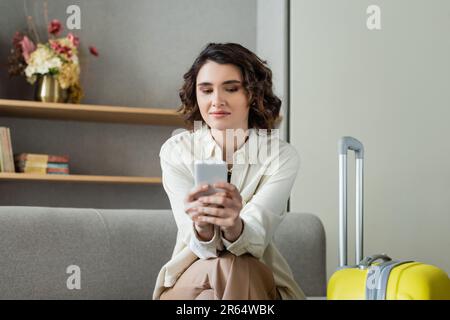 giovane donna sorridente con capelli brunette utilizzando smartphone mentre si siede sul divano vicino alla borsa da viaggio, scaffali con libri e vaso con fiori su blozzed ba Foto Stock