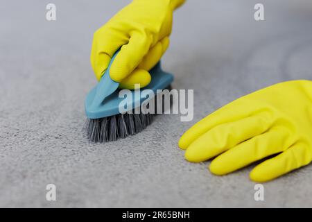 primo piano delle mani in guanti di gomma gialla pulizia tappeto grigio con spazzola Foto Stock