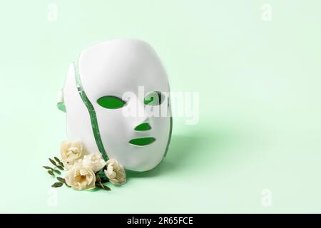 Maschera cosmetica a LED trattamento rigenerativo su sfondo verde con delicati fiori di rosa e un posto per il tuo testo. Salute e bellezza della f Foto Stock