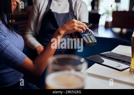 Giovane donna in abbigliamento casual che paga per la cena con la macchina da carte accanto alla cameriera Foto Stock