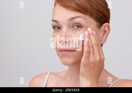 Ritratto di caucasico corto faccia donna di mezza età con lenticchi e capelli rossastri applicando crema sul viso su sfondo bianco guardando la macchina fotografica Foto Stock