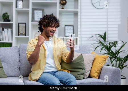 Gioioso e felice giovane uomo seduto sul divano da solo a casa, ispanico celebrando la vittoria e il successo, ha ottenuto buone notizie utilizzando app sul telefono. Foto Stock
