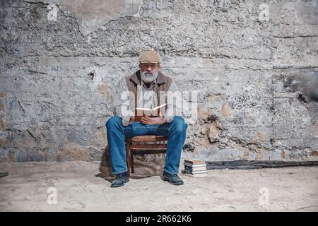 L'uomo con barba grigia si siede su una sedia contro una parete di cemento, leggendo un libro Foto Stock