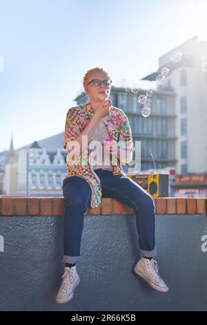 Giovane uomo che soffia bolle sul soleggiato balcone della città Foto Stock