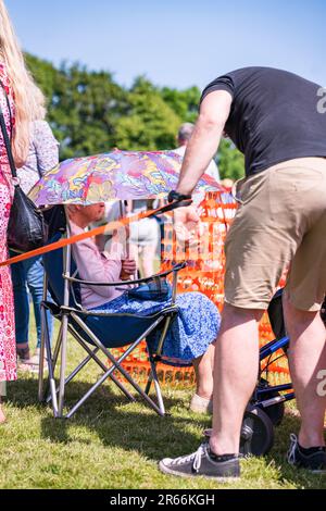 Donna anziana che indossa un abito estivo blu seduto su una sedia pieghevole che tiene un ombrello come una tenda da sole. Foto Stock
