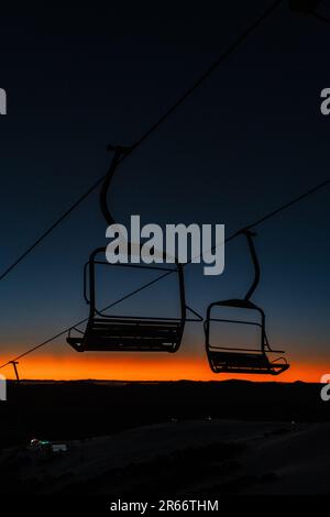 Una vista panoramica di due impianti di risalita in cima alla collina innevata al tramonto, con le maestose montagne sullo sfondo Foto Stock