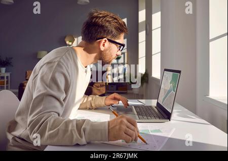 Uomo che usa il suo computer portatile mentre lavora con mappe catastali digitali e cartacee della città Foto Stock