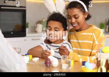 Madre afro-americana felice ed il suo figlio carino che dipinge le uova di Pasqua al tavolo in cucina Foto Stock