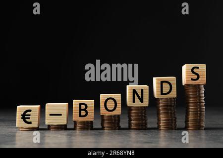 Monete impilate e Eurobond di parole in cubetti di legno con lettere sul tavolo grigio Foto Stock