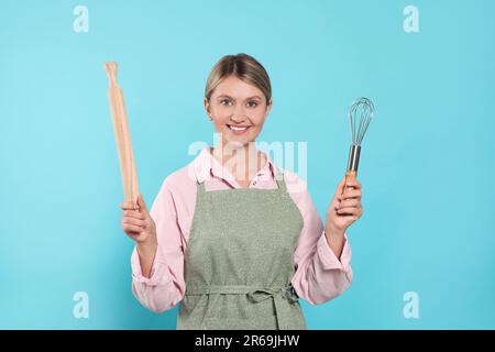 Bella giovane donna in grembiule pulito con utensili da cucina su sfondo azzurro Foto Stock