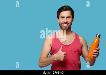 Uomo con bottiglia termica arancione che mostra i pollici su sfondo azzurro. Spazio per il testo Foto Stock