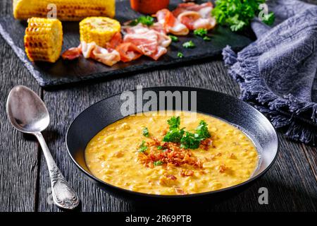 zuppa di mais cremosa con pancetta fritta e prezzemolo sbriciolati in ciotola nera su un tavolo di legno scuro, con mais alla griglia in pannocchie e baco affumicato Foto Stock