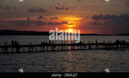 Gruppo di persone non riconoscibili in silhouette seduta sul Molo Jetty di guardare il tramonto tramonto sul lago Steinhuder Meer in Germania Foto Stock