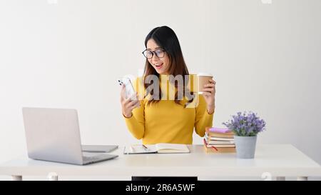 Una giovane studentessa asiatica allegra ed eccitata o un dipendente sta guardando un messaggio incredibile e inaspettato sul suo telefono mentre sorseggia un caffè Foto Stock